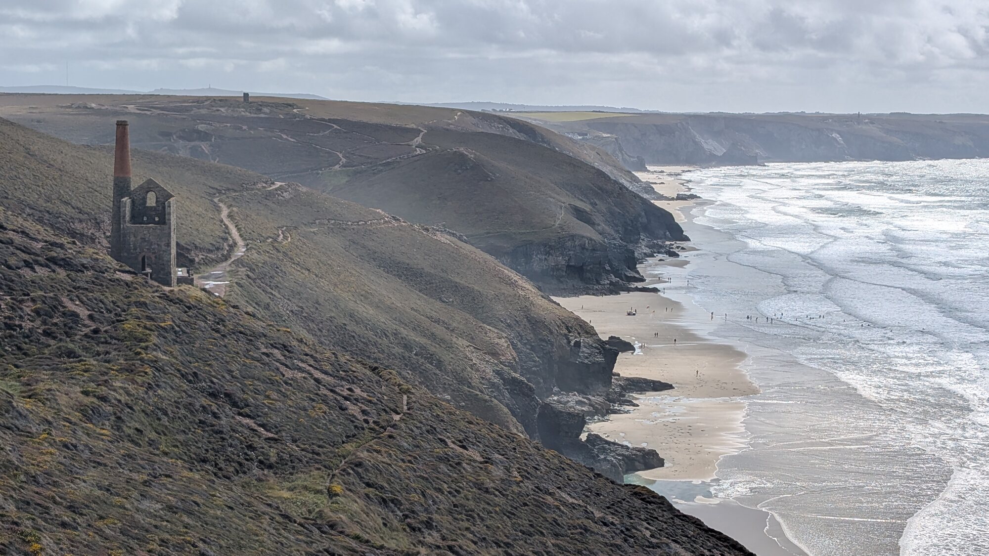 The Wheal Coates engine house stands on a cliff above Chapel Porth on Cornwall's Tin Coast, with surf-curled bay below and the wider mining landscape of cliffs and engine-house ruins beyond.