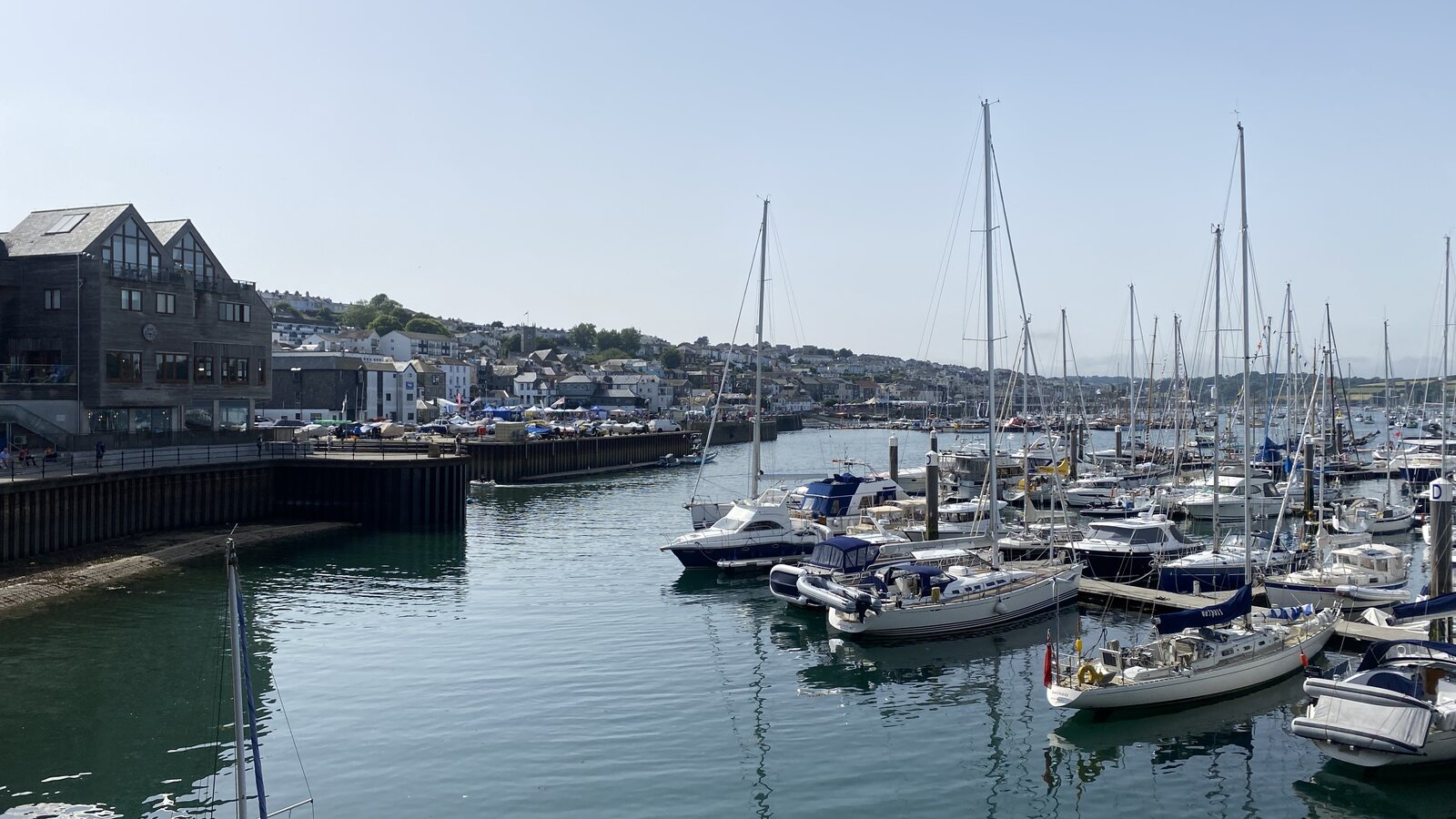 The marina pontoons at Falmouth's Custom House Quay area on a clear summer day: white-hulled sailing yachts and motorboats moored at the floating jetties, the harbour-front buildings of central Falmouth rising on the hillside in the background, and marquees and a crowd visible on the quayside in the middle distance during what appears to be Falmouth Week.