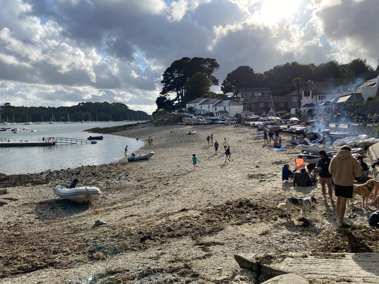 Helford Passage village beach with dinghies pulled up on the sand, families on the beach, sailboats in the river behind, and the village cottages along the waterfront.