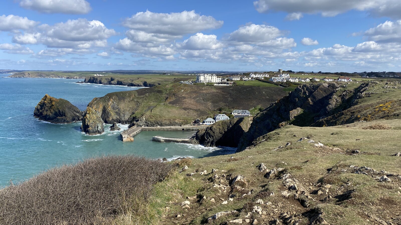 Mullion Cove on the western Lizard, with stone harbour walls, a sandy beach, and dramatic rocky outcrops behind. A small fishing fleet shelters inside the harbour.