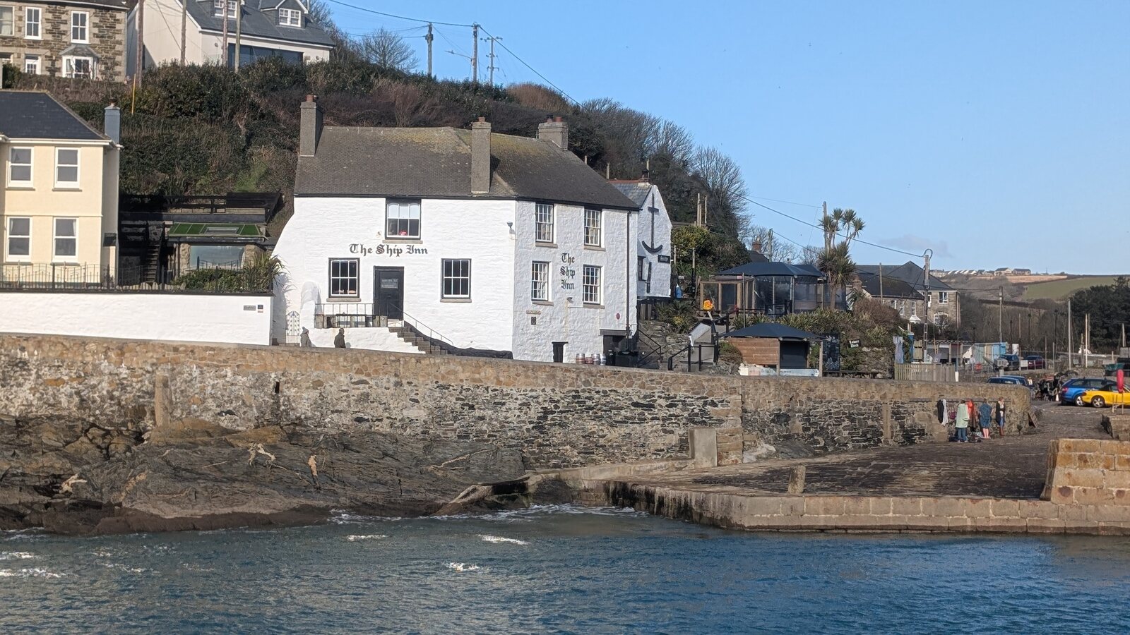 The white-painted Ship Inn above the harbour wall at Porthleven, with its name painted on the walls, stone steps up to the door and the sea in the foreground.
