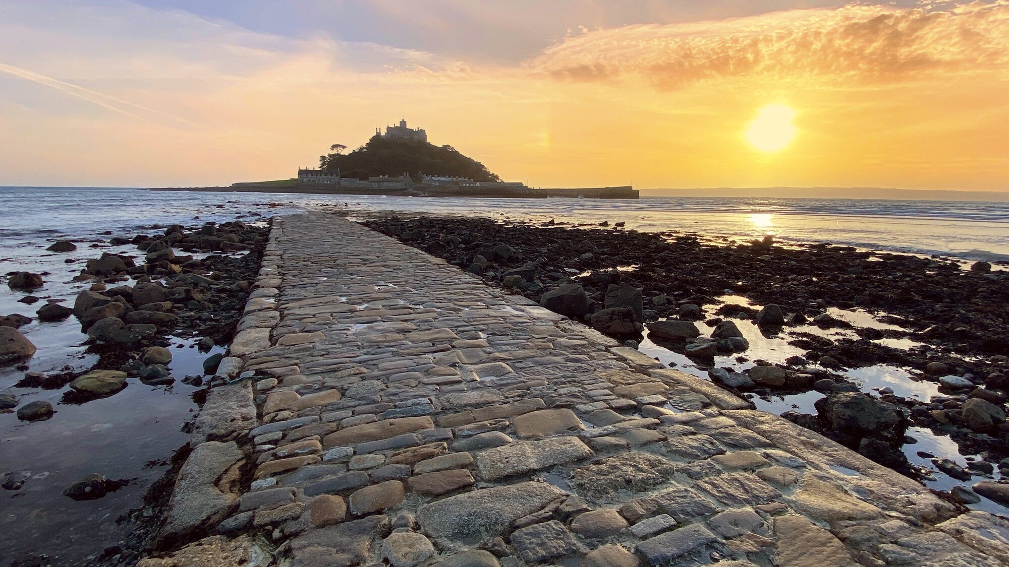 St Michaels Mount silhouetted against an orange sunset sky, with the cobbled granite causeway stretching across the seabed at low tide and kelp-strewn rocks and wet sand on either side.