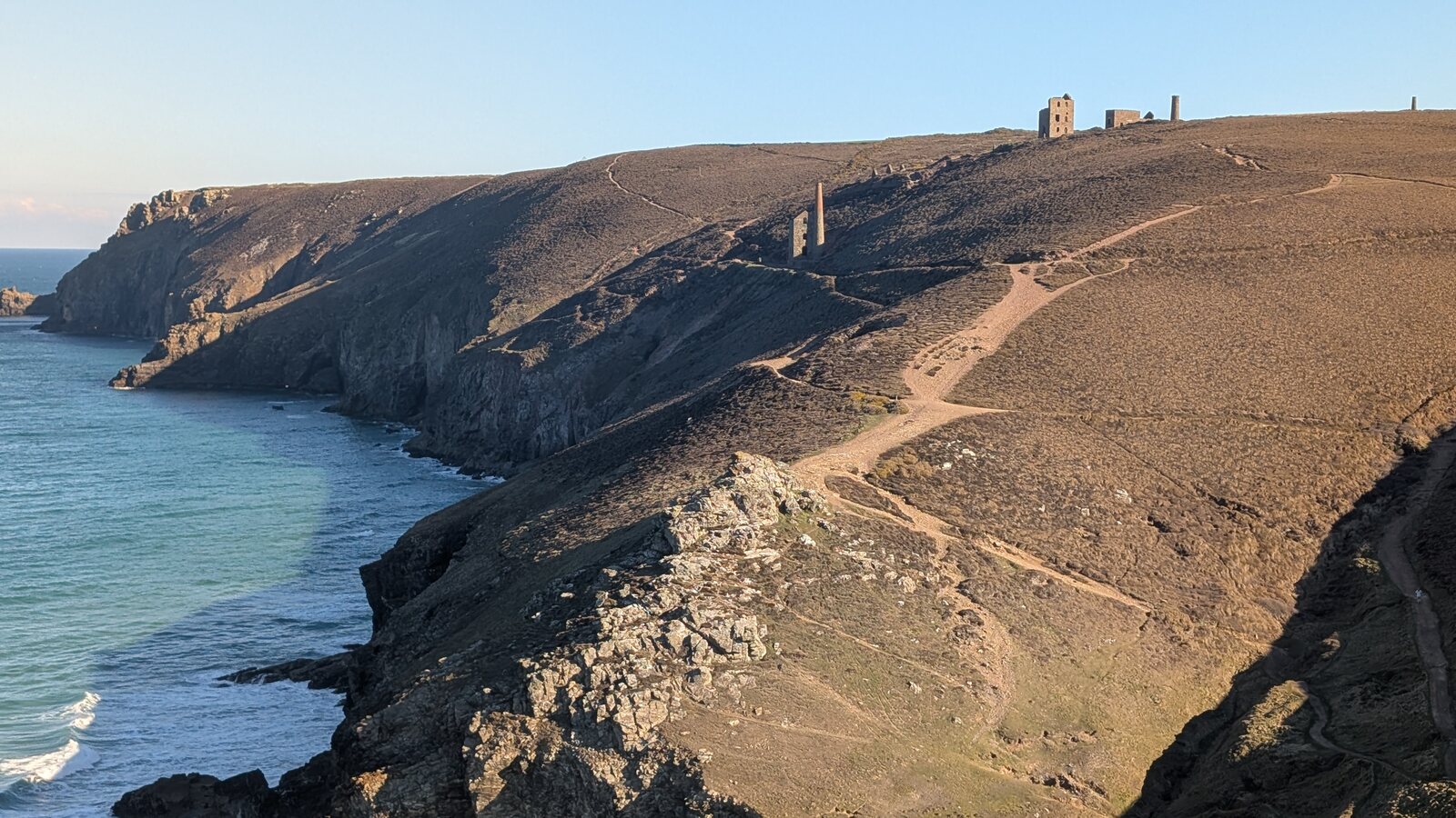 The Wheal Coates engine houses on the skyline above the cliffs at Chapel Porth, with the coast path climbing the brown heath in low evening sun and the sea below.