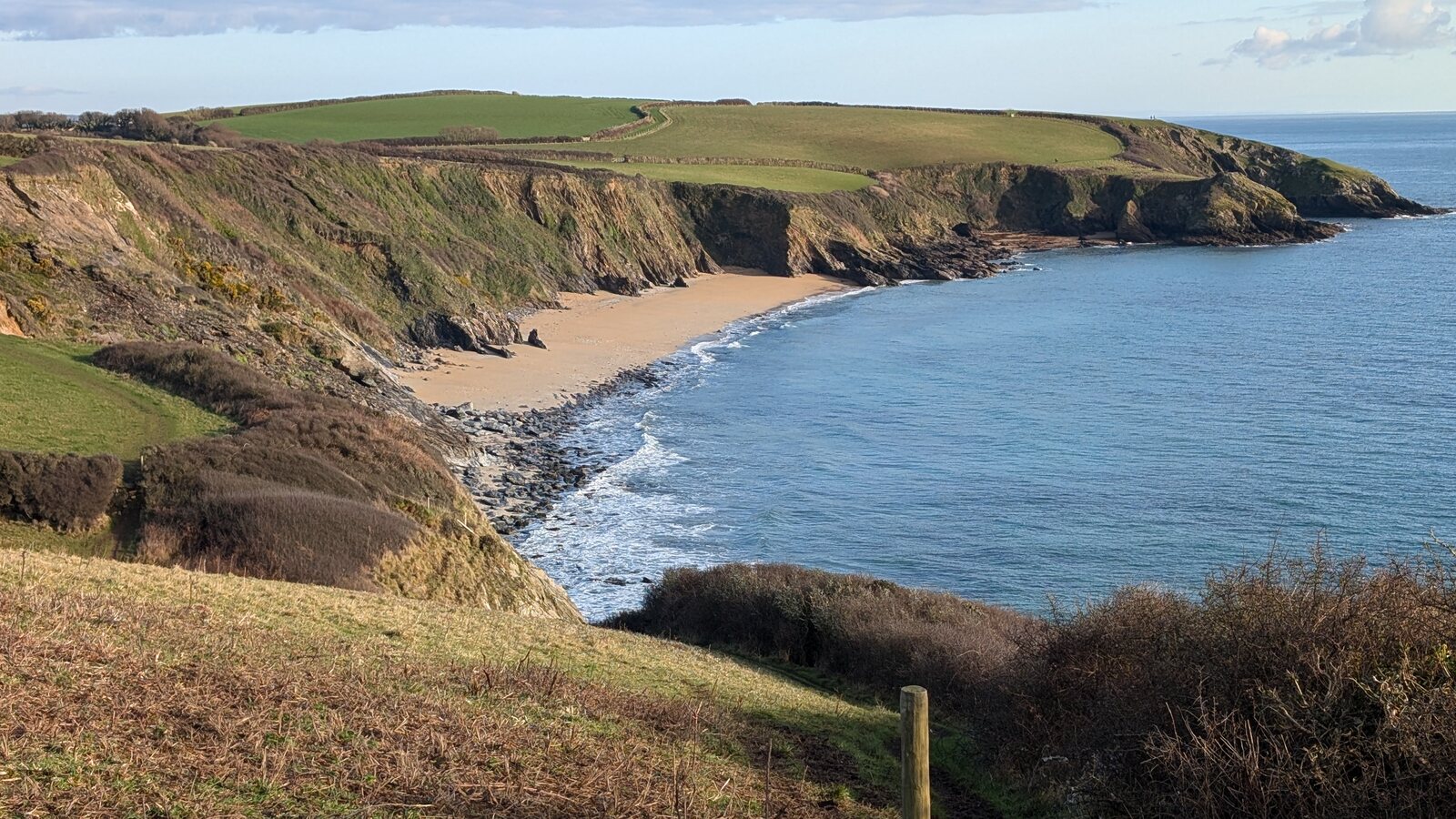 Porthbeor Beach from the coast path: a curve of golden sand below steep green cliffs, with waves breaking on the rocks and farmland running out to the headland.
