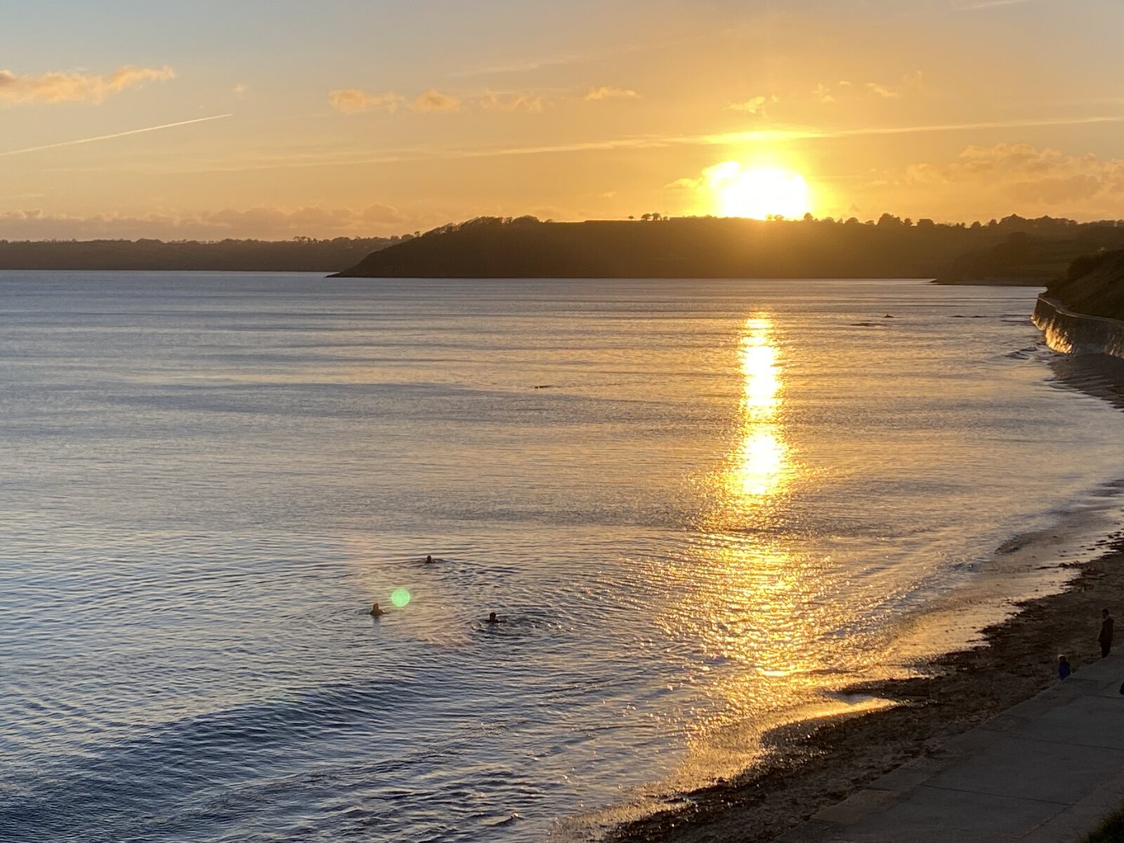 Wild swimmers in calm water at Castle Beach, Falmouth, with the sun rising over a distant headland and a golden trail of light reflected on the surface of Falmouth Bay.