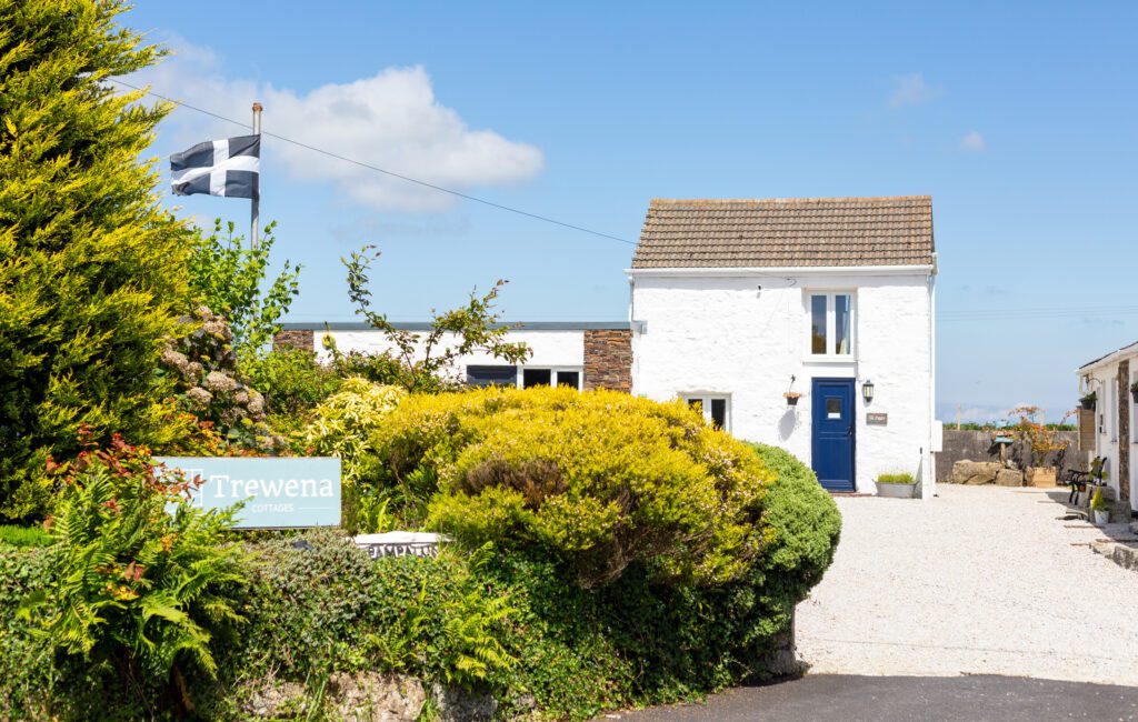 Exterior view of Trewena Cottages with the Cornish flag flying, surrounded by vibrant greenery and a blue sky.
