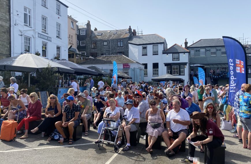 A lively crowd gathers in Falmouth during the International Sea Shanty Festival, sitting and socializing near the Quayside Inn with festival banners and historic buildings in the background.