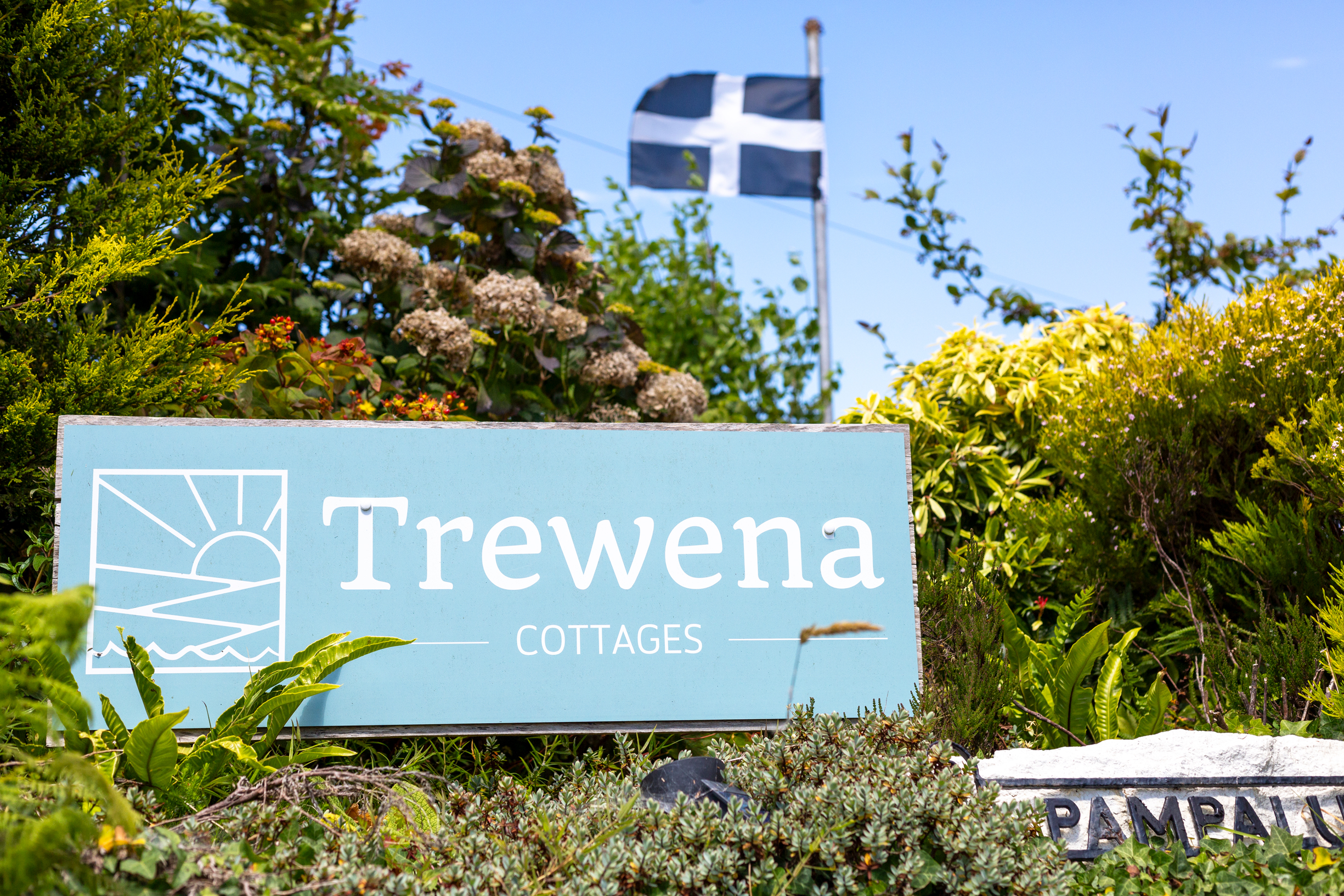 Trewena Cottages sign with a Cornish flag flying in the background, surrounded by vibrant greenery.