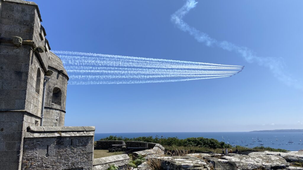 The Red Arrows flying in formation above Pendennis Castle in Falmouth, with smoke trails against a clear blue sky and boats gathered in the bay below.