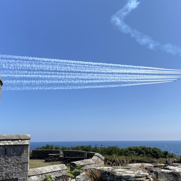 The Red Arrows flying in formation above Pendennis Castle in Falmouth, with smoke trails against a clear blue sky and boats gathered in the bay below.