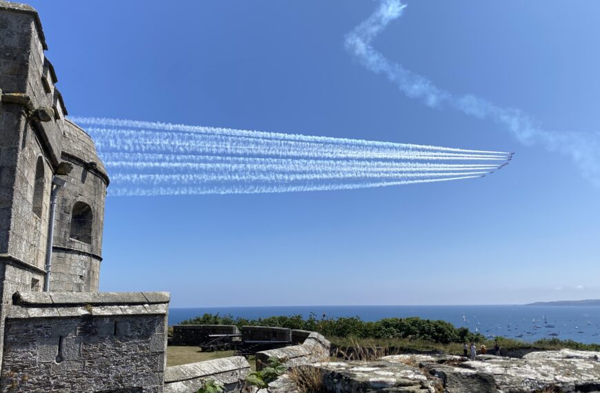The Red Arrows flying in formation above Pendennis Castle in Falmouth, with smoke trails against a clear blue sky and boats gathered in the bay below.