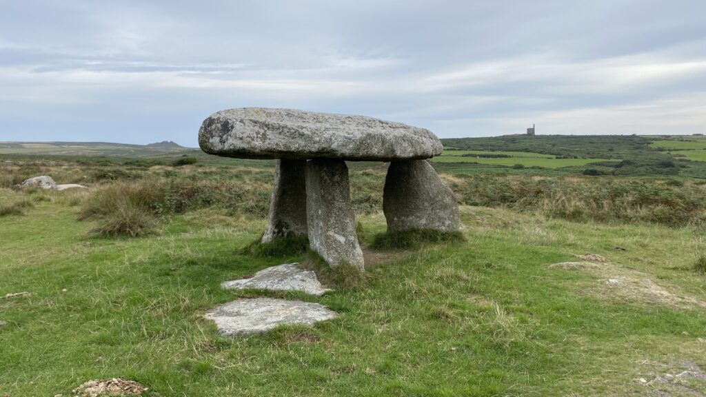 Lanyon Quoit, a Neolithic dolmen on the Penwith Moors in Cornwall, standing under a cloudy sky with green fields and an old mine chimney in the background.