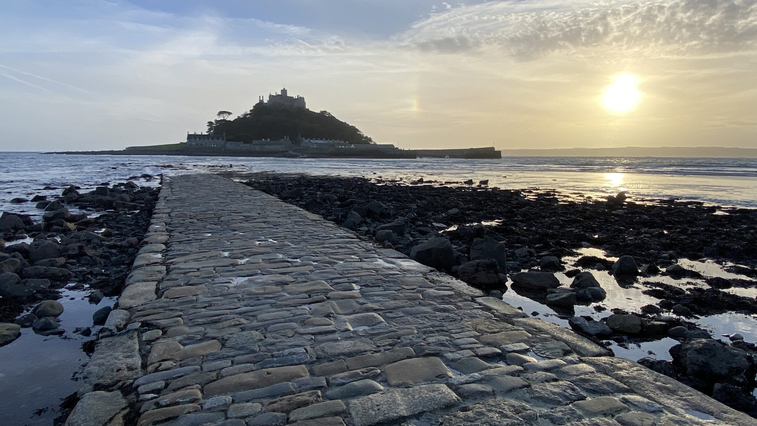Cobblestone causeway leading to St Michael’s Mount in Cornwall at sunset, with the tide out and golden light on the water.