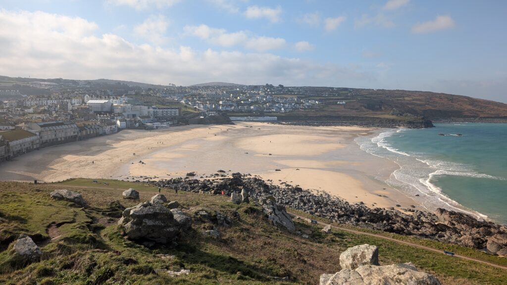 View of Porthmeor Beach and the town of St Ives in Cornwall from the clifftop coast path, with golden sand, turquoise sea and scattered walkers.