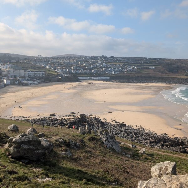 View of Porthmeor Beach and the town of St Ives in Cornwall from the clifftop coast path, with golden sand, turquoise sea and scattered walkers.