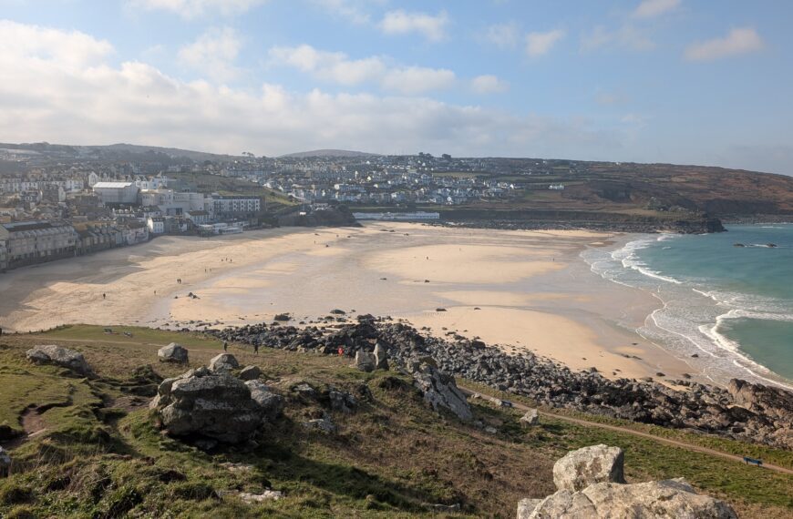 View of Porthmeor Beach and the town of St Ives in Cornwall from the clifftop coast path, with golden sand, turquoise sea and scattered walkers.