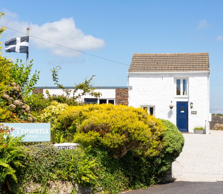 Exterior view of Trewena Cottages with the Cornish flag flying, surrounded by vibrant greenery and a blue sky.
